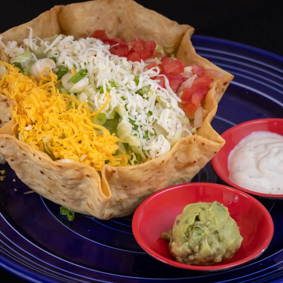 A taco salad in a crispy tortilla bowl with shredded cheese, lettuce, tomatoes, and sides of guacamole and sour cream.