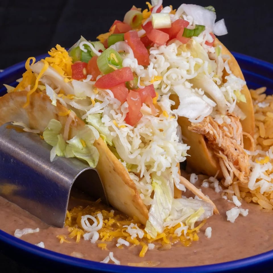 Two crispy chicken tacos with lettuce, cheese, tomatoes, and onions, served with rice and refried beans.